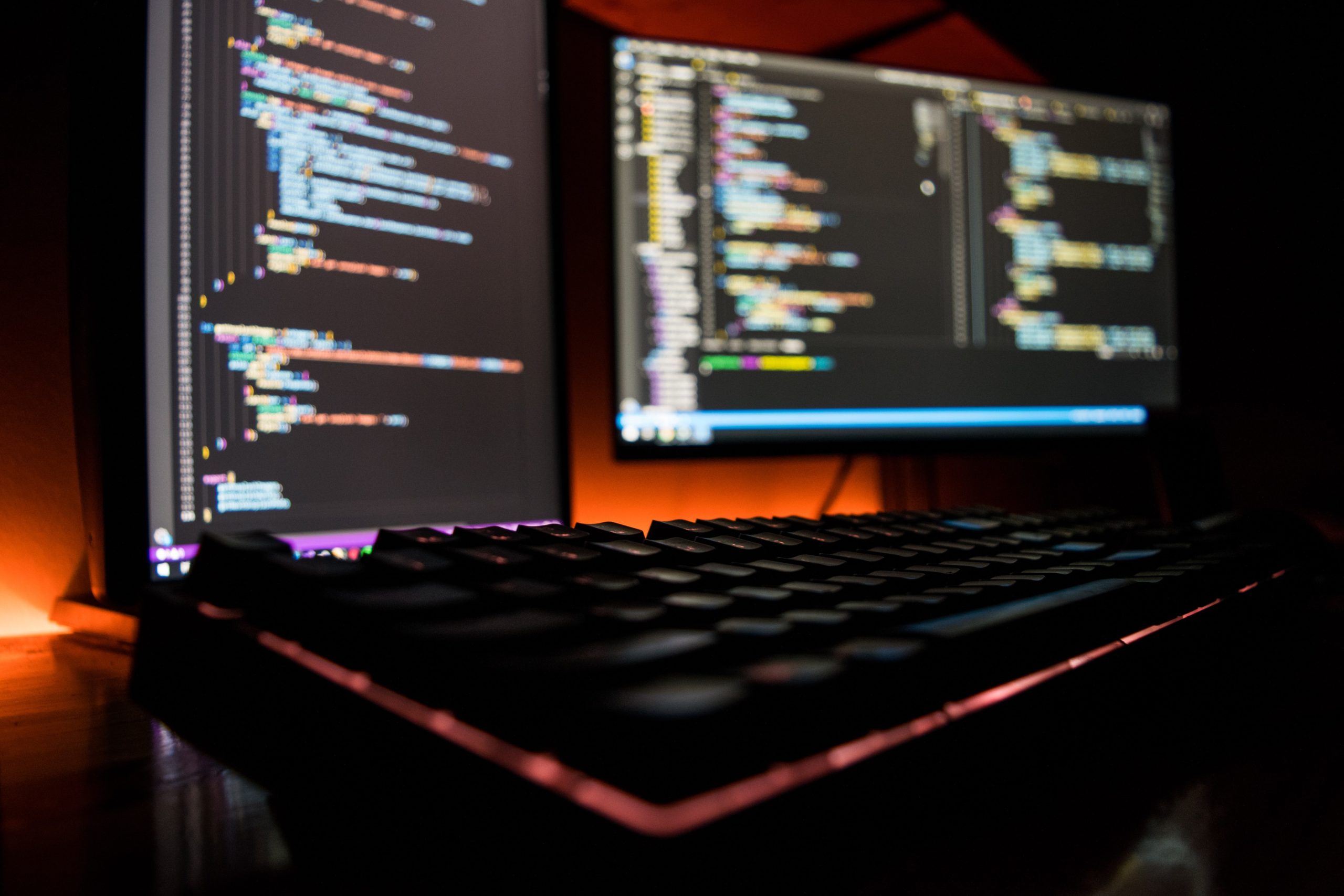 Black keyboard and monitors in soft focus with multicolored html code in front of an orange backlit background