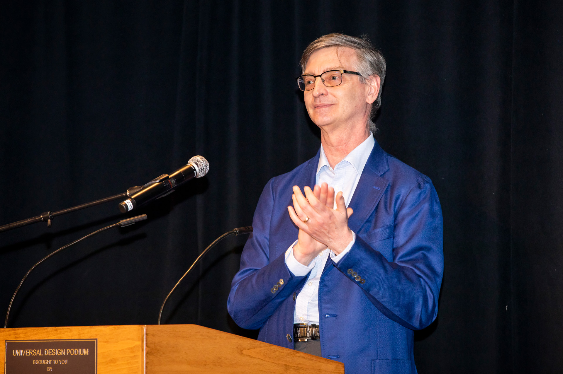 Photograph of Bill Gropp in a blue jacket applauding and smiling at a podium; he is standing in front of a black back-drop.